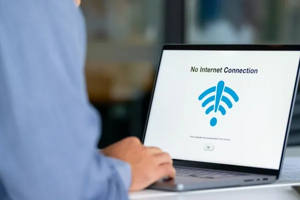 A person wearing a blue shirt is viewed from over the shoulder as they look at a laptop screen displaying a "No Internet Connection" message with a blue Wi-Fi symbol.